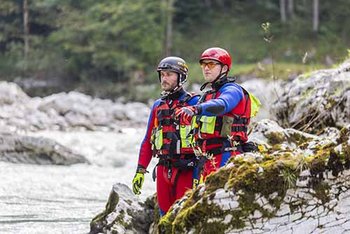 Zwei Strömungsretter stehen mit Helm und blau-roter Einsatzkleidung auf Felsen an fließendem Gewässer